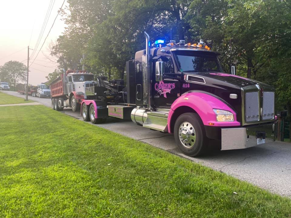 A black and pink tow truck with flashing blue lights pulling a second truck on a tree-lined road.
