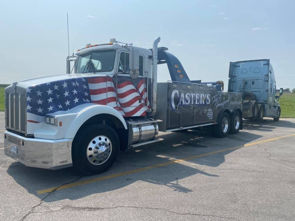 A tow truck with an American flag design is towing a semi-truck on a sunny day. The tow truck has 