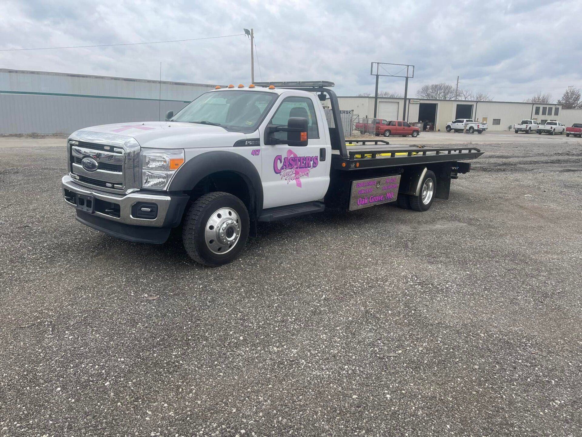 A white tow truck with pink accents, parked on a gravel lot, under an overcast sky.