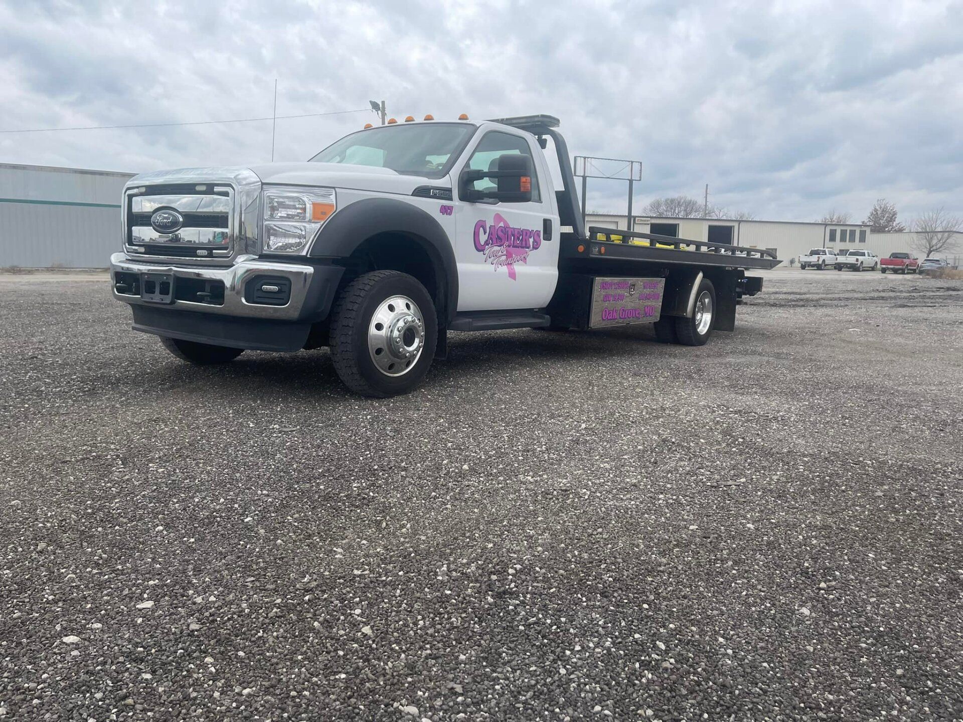 White tow truck with a flatbed in a gravel parking lot on a cloudy day. The truck has pink lettering on the door.