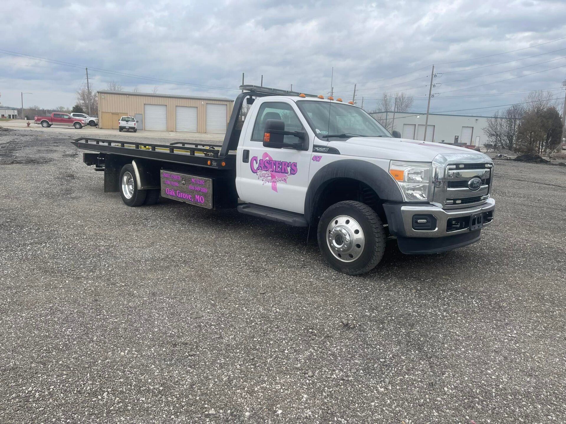 A white tow truck with pink lettering is parked on a gravel lot in front of a beige building under an overcast sky.