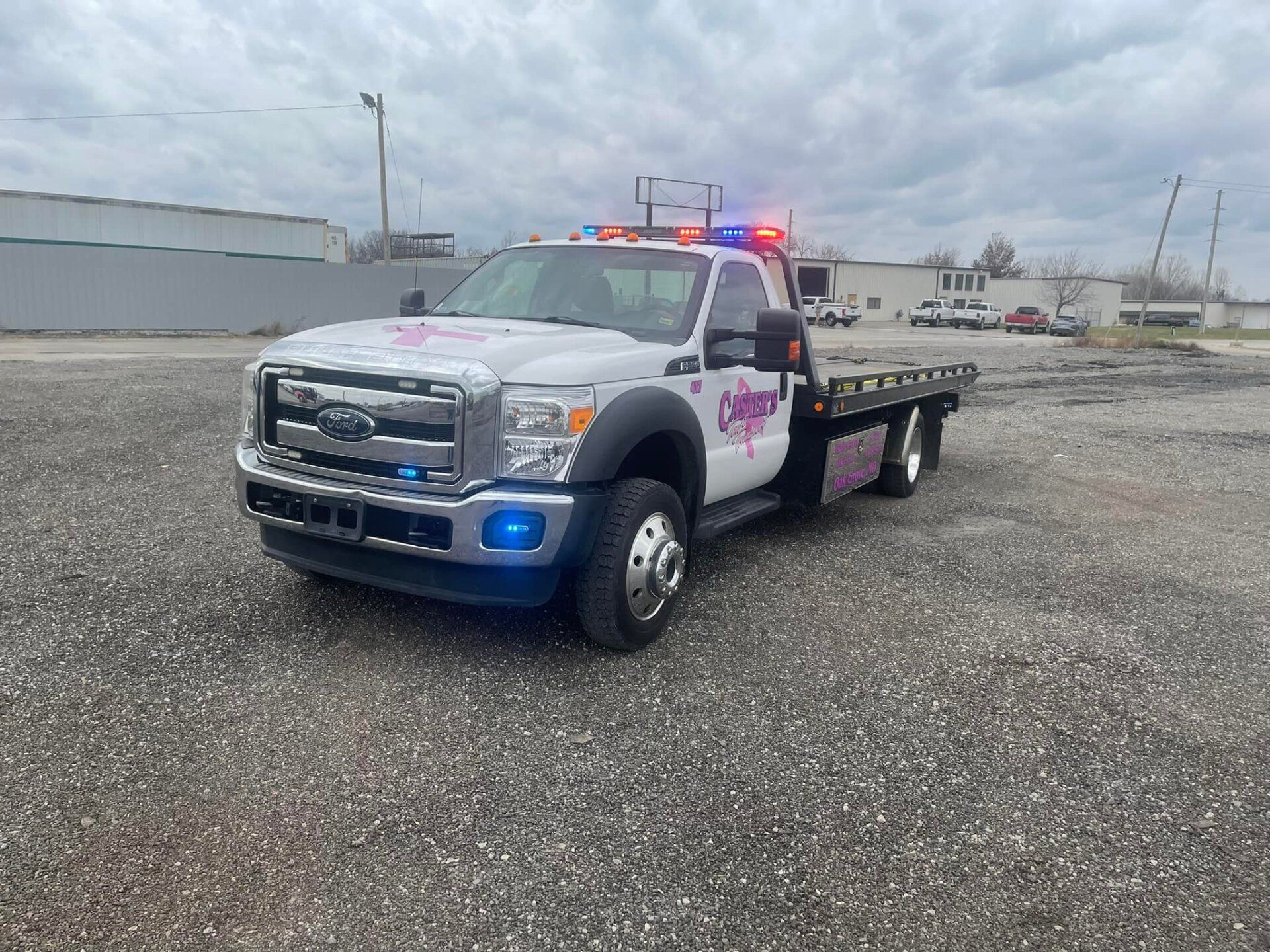 White tow truck with pink accents and flashing lights parked on gravel, outdoors under cloudy skies.