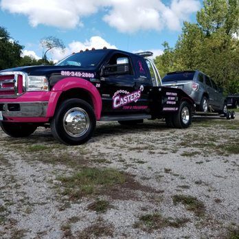A black and pink tow truck with a gray SUV on its flatbed, parked on gravel under a cloudy sky.