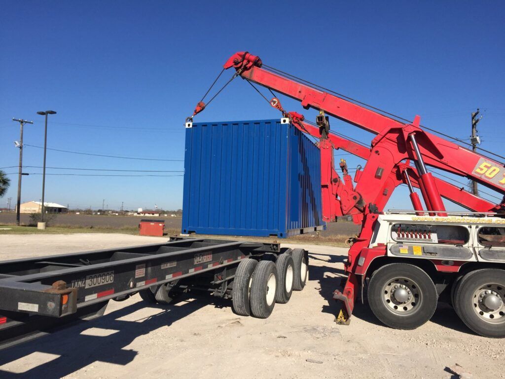 A blue shipping container being lifted by a red tow truck onto a trailer, outdoors.