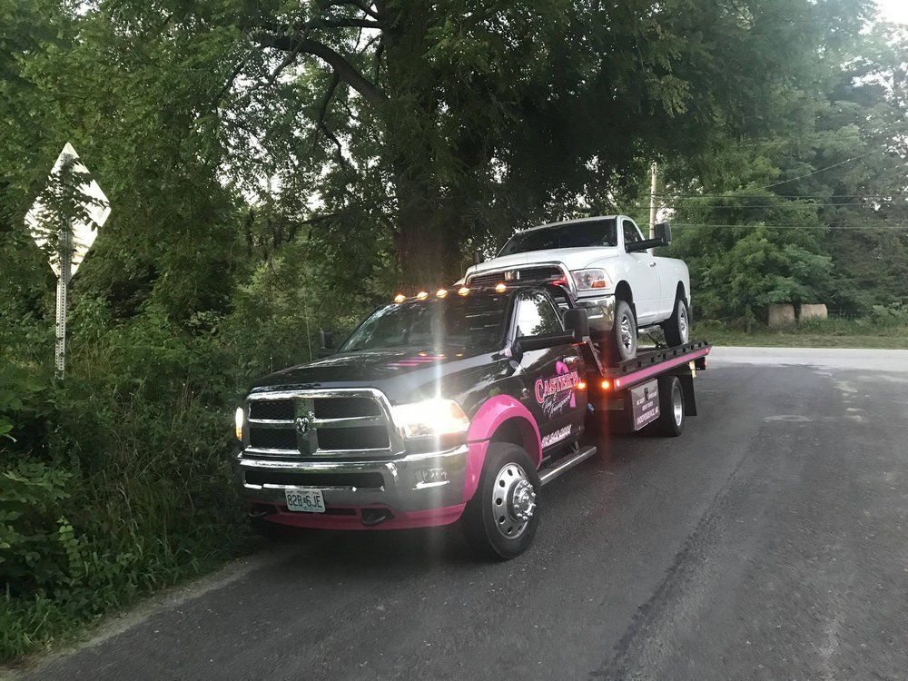 A black tow truck with pink accents carries a white pickup truck down a road, near green foliage.