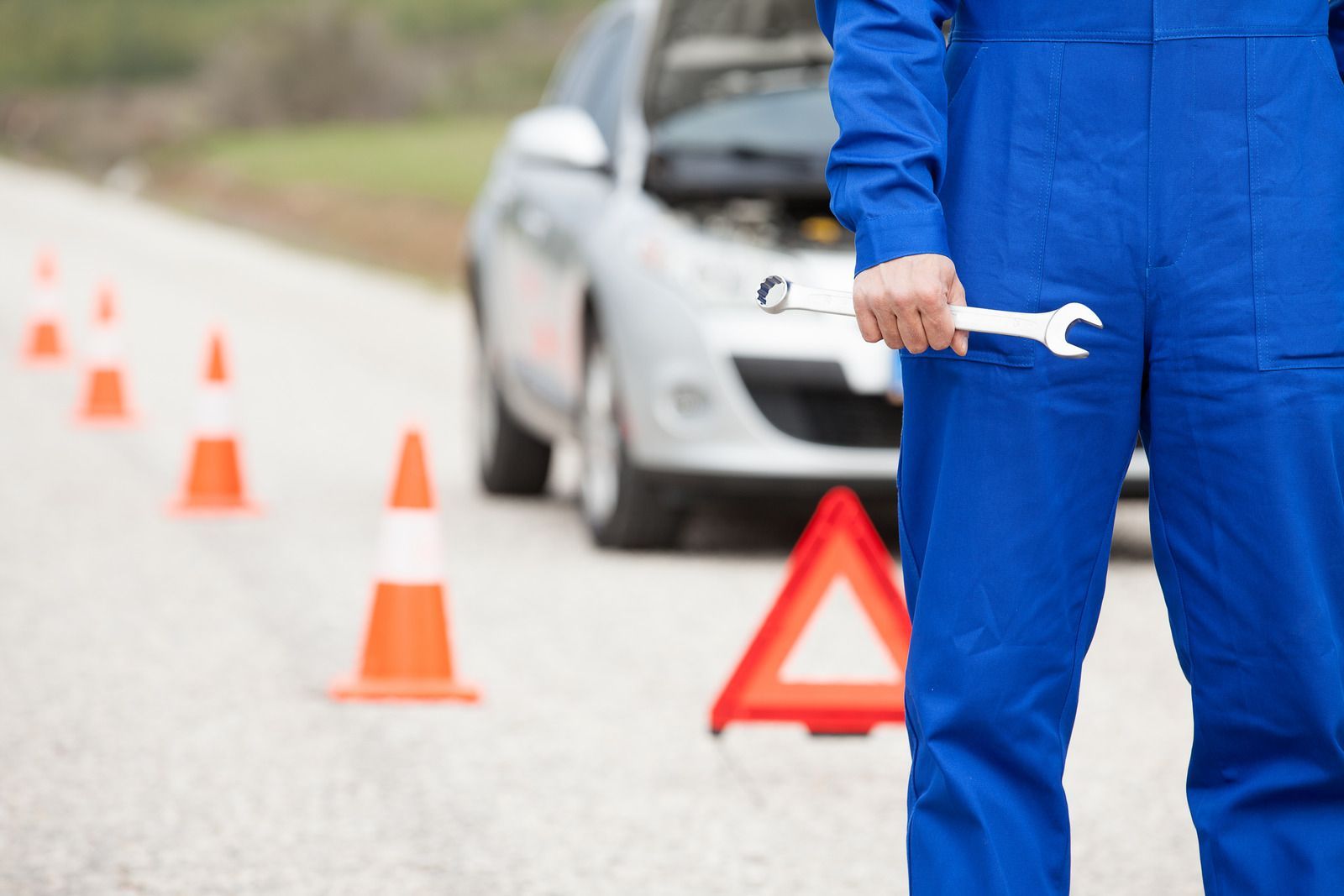Mechanic in blue overalls holding a wrench, standing near a disabled silver car on a roadside.