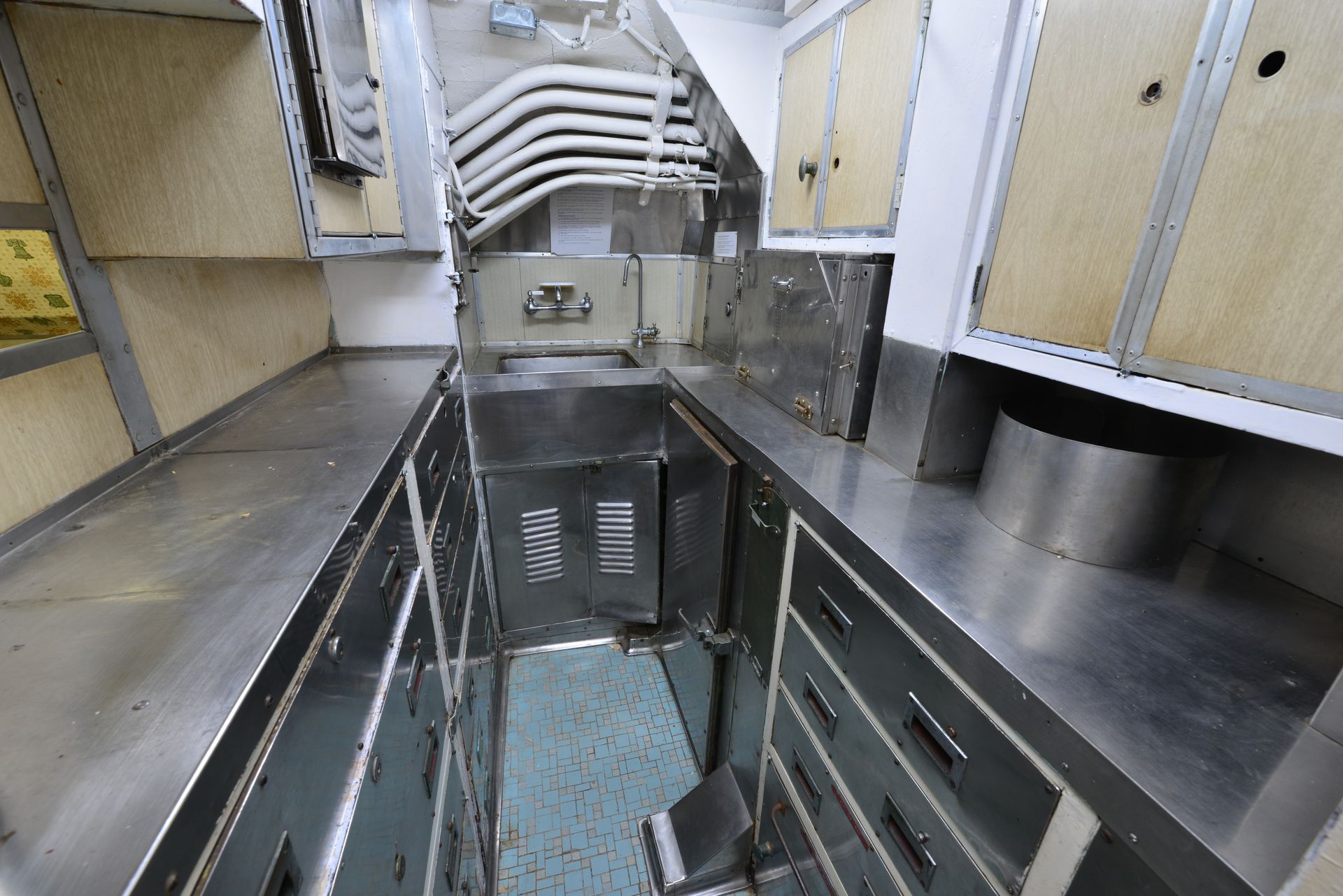 Interior of a narrow, industrial kitchen with stainless steel counters, cabinets, and ventilation pipes.