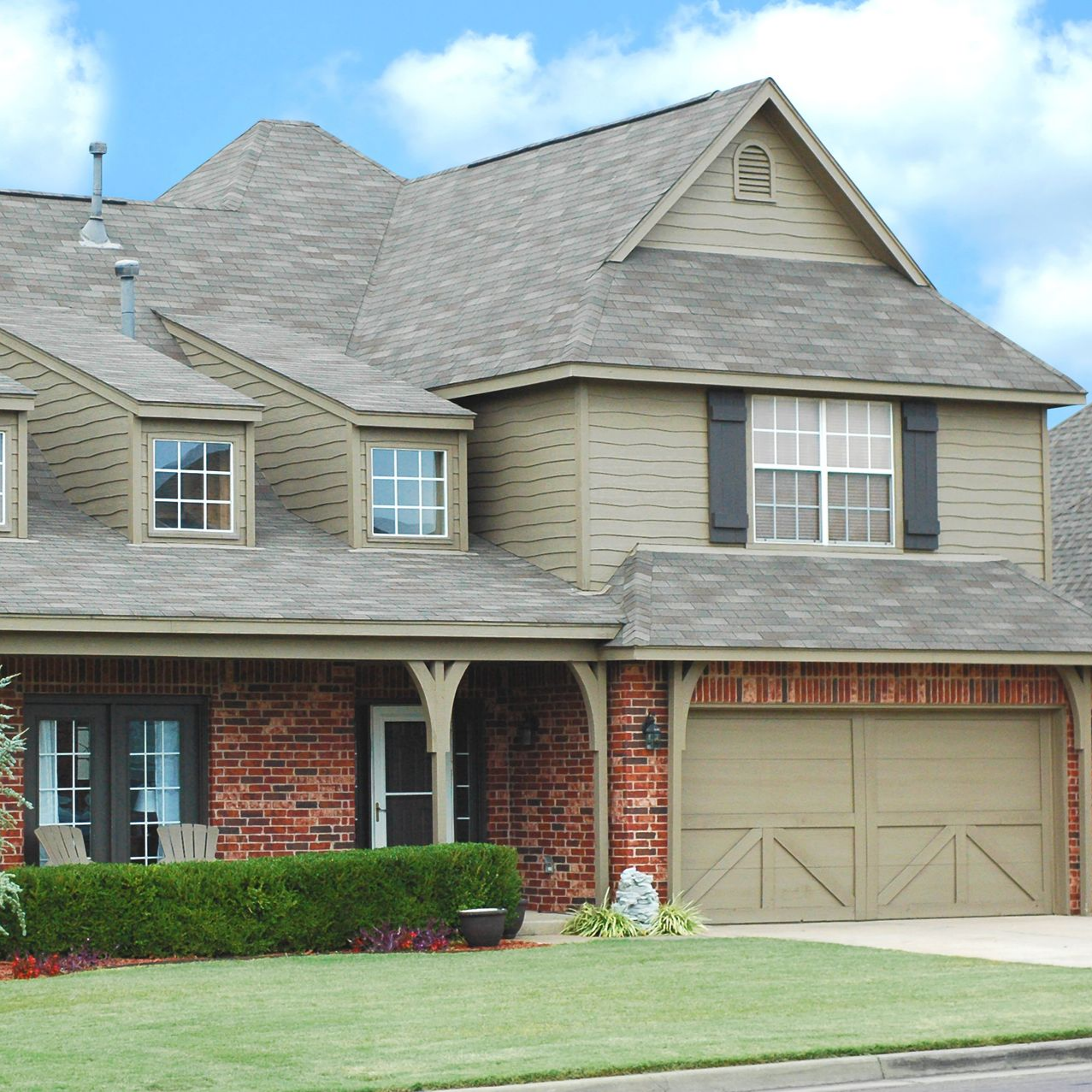 Two-story house with tan siding, red brick, gray roof, and a beige garage door, set in front of a green lawn.
