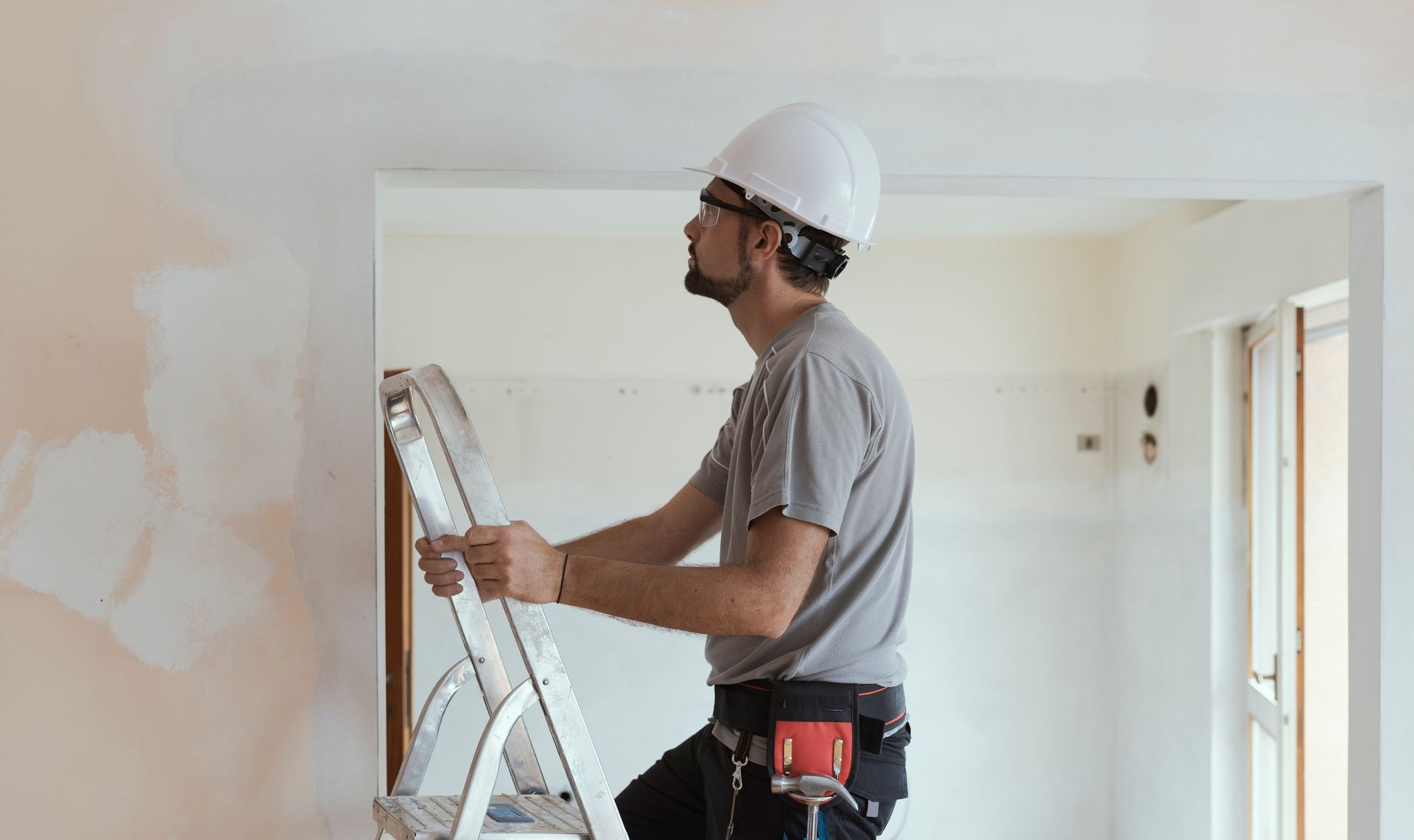 Construction worker on a ladder inspecting ceiling, wearing hard hat and tool belt. Interior setting with repaired walls.