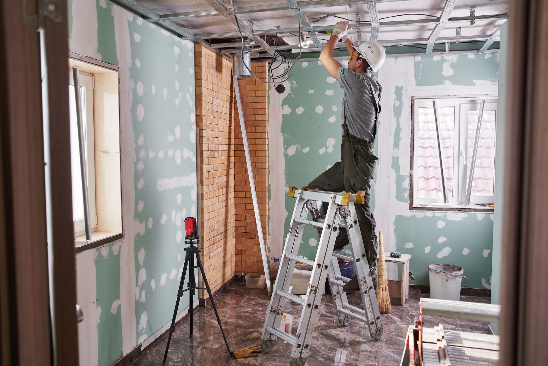 Person in a hard hat on a ladder, working on ceiling wiring in a room under construction with drywall and windows.