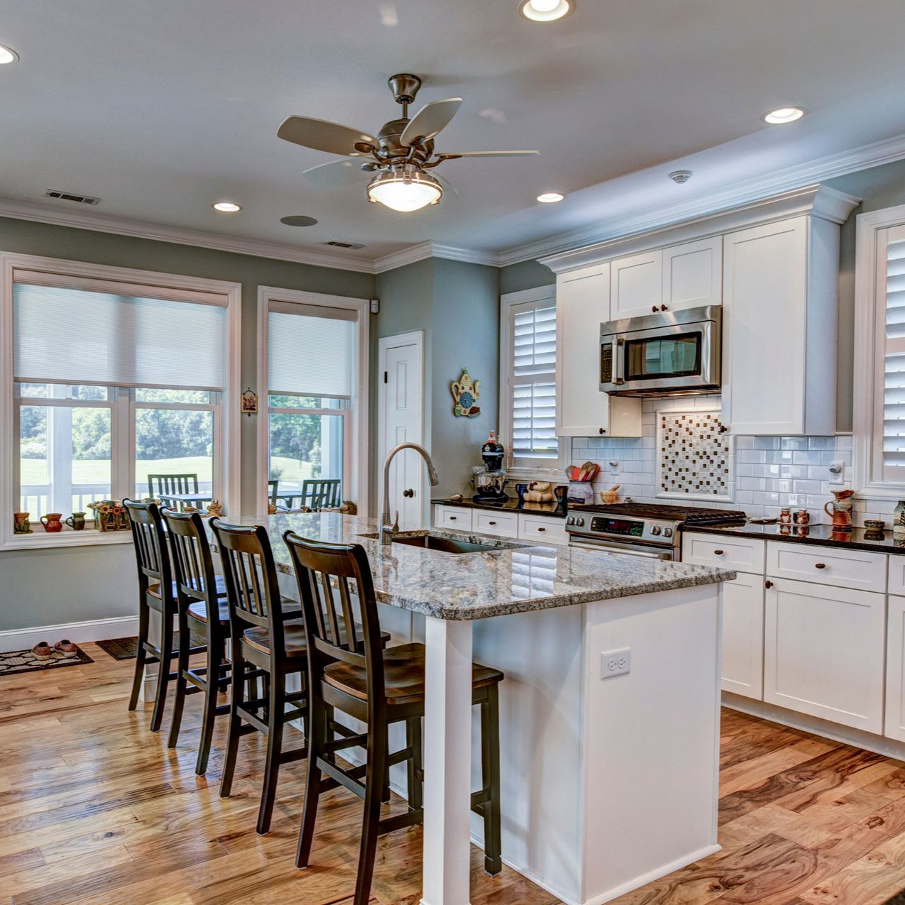 Kitchen with white cabinets, island, and wooden floor. Window with blinds, chairs, and ceiling fan.