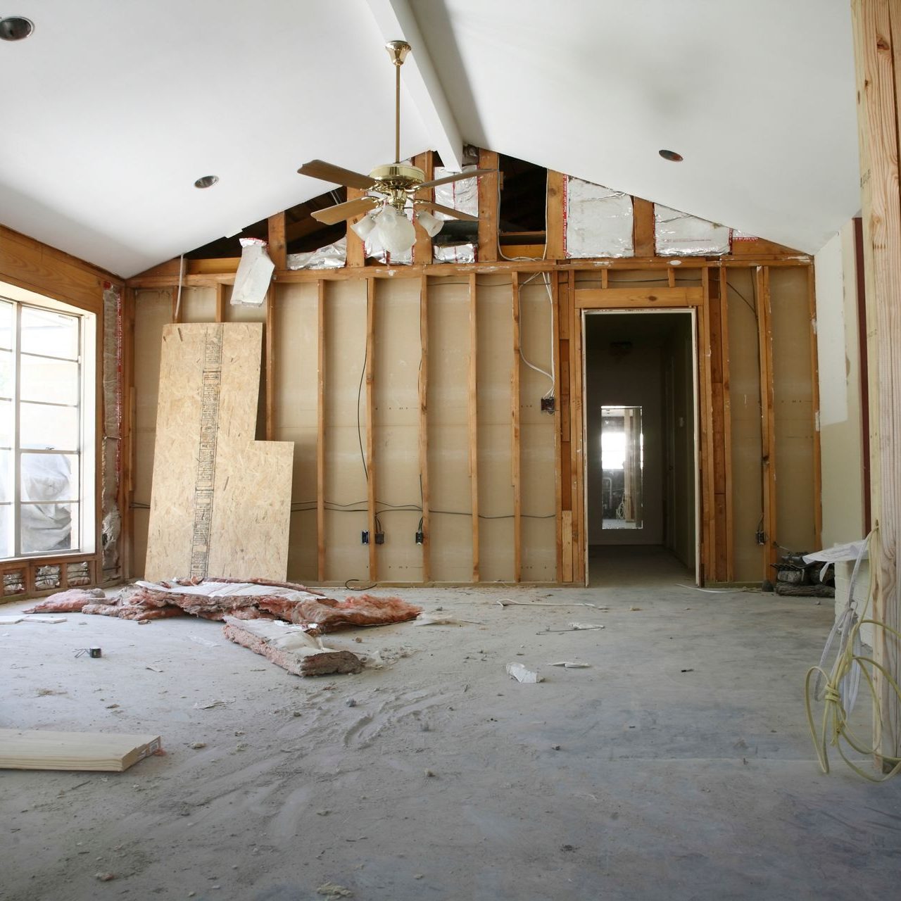 Interior room undergoing renovation, exposed wood framing, ceiling fan, concrete floor, doorway.