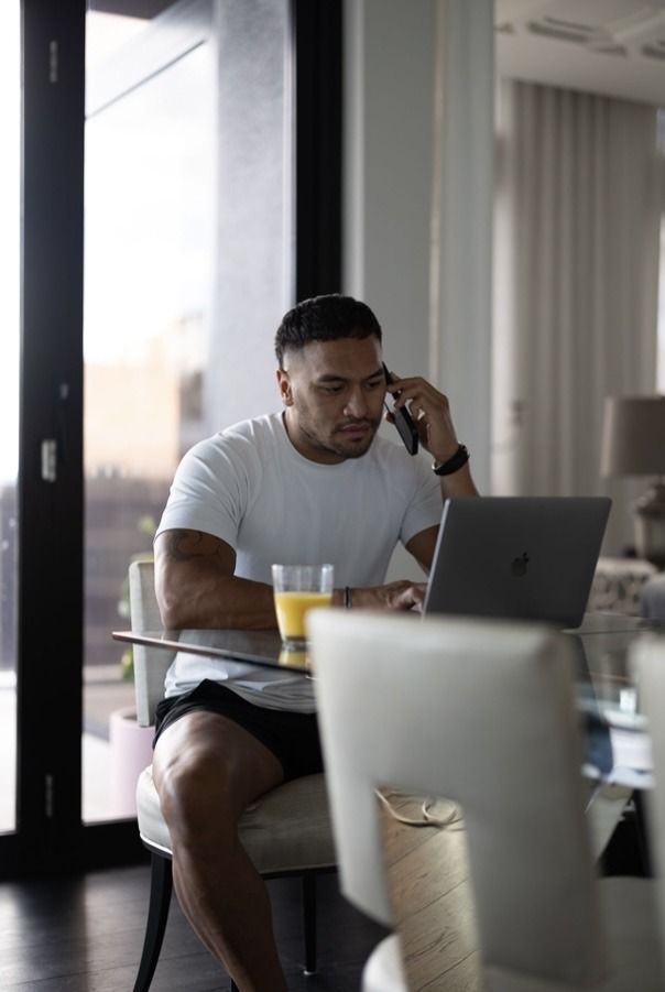 A man is sitting at a table using a laptop and talking on a cell phone.