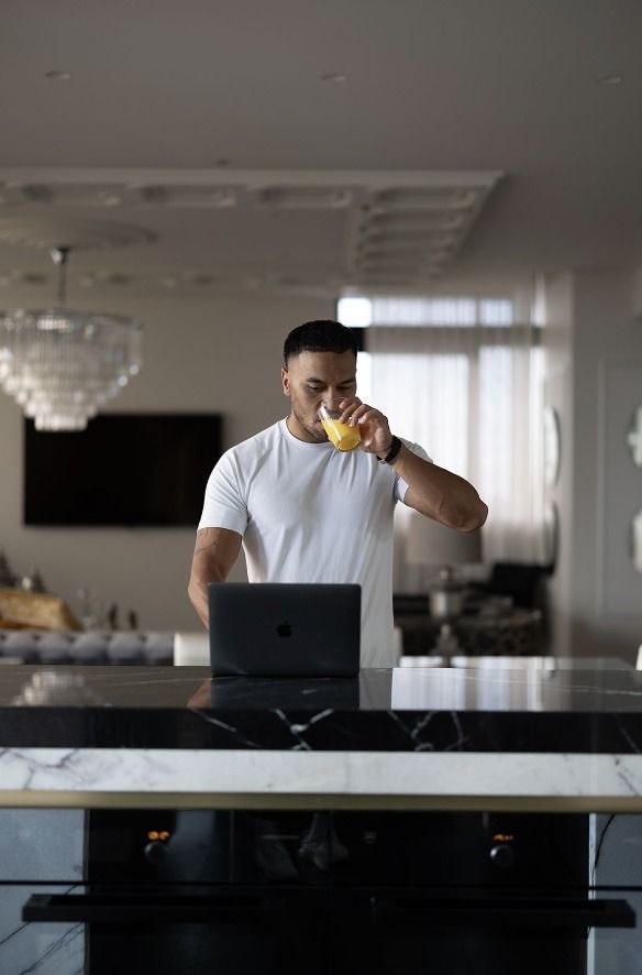 A man is drinking orange juice while using a laptop computer in a kitchen.