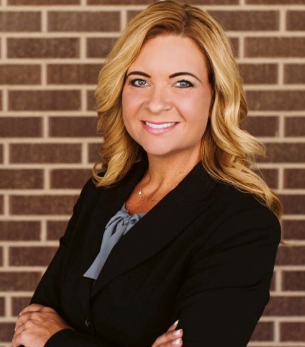 Woman with blonde hair wearing a black blazer smiles, arms crossed, standing in front of a brick wall.