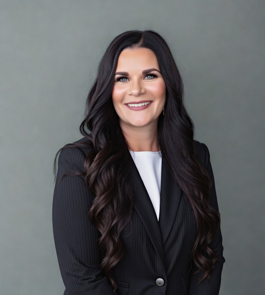 Woman with dark hair, wearing a black blazer over a white top, smiling in front of a gray backdrop.