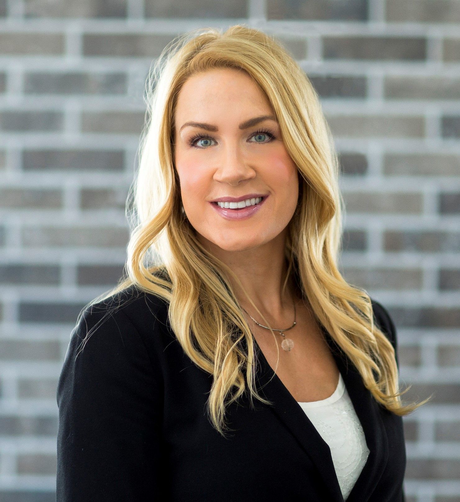 Blonde woman in a black blazer, smiling, in front of a brick wall.