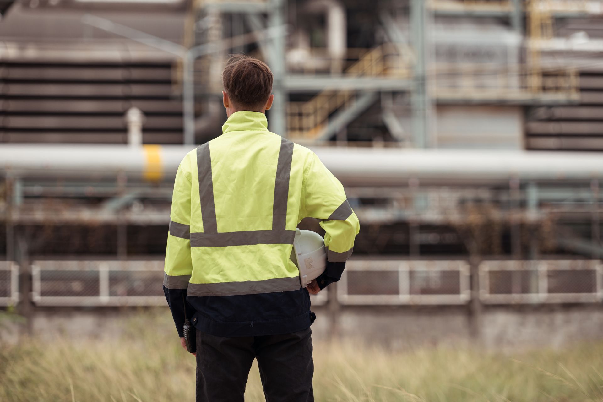 Person in high-visibility jacket looking at industrial facility; holding a white hard hat.