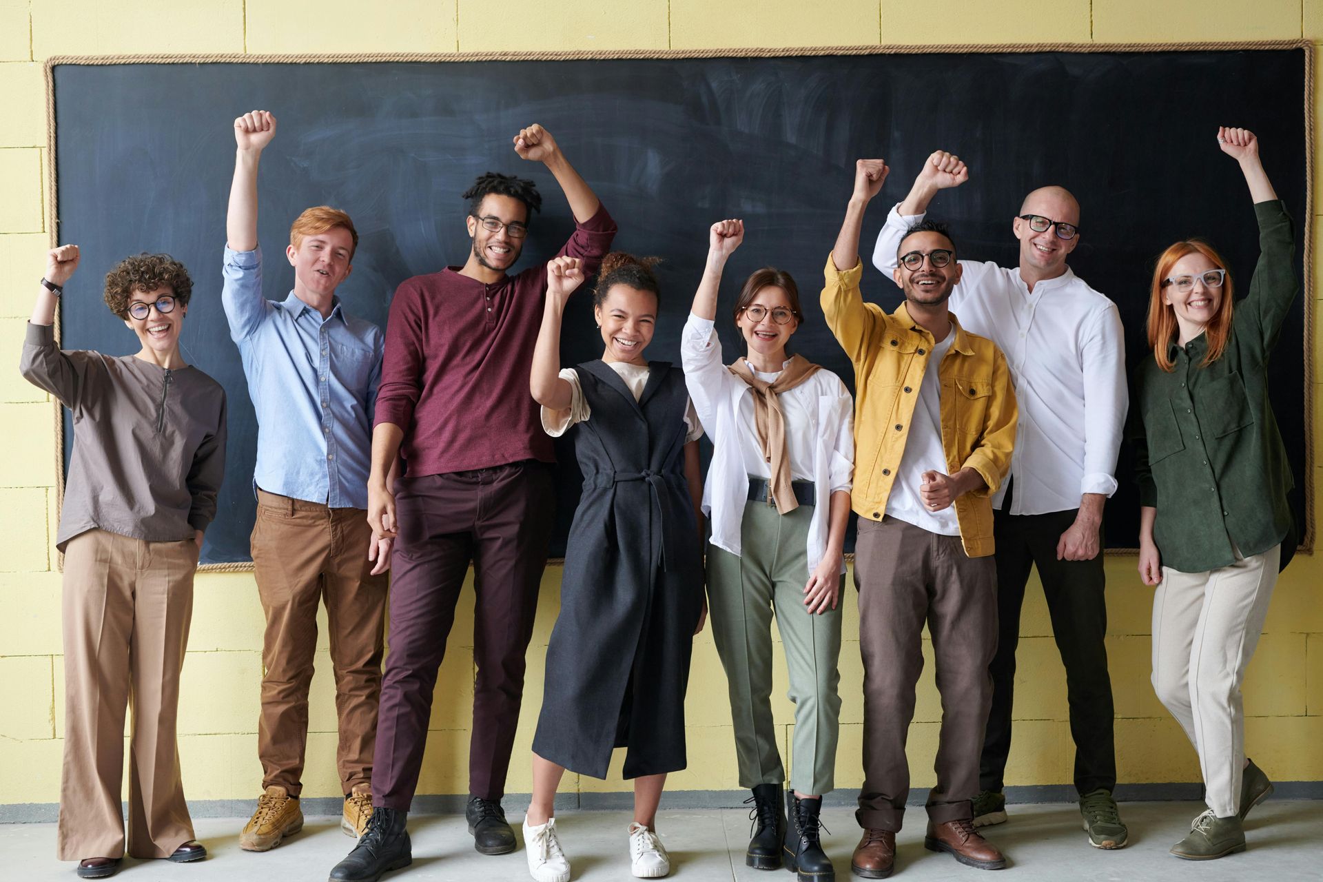 Seven people standing in a row, smiling and raising their fists in front of a blackboard.