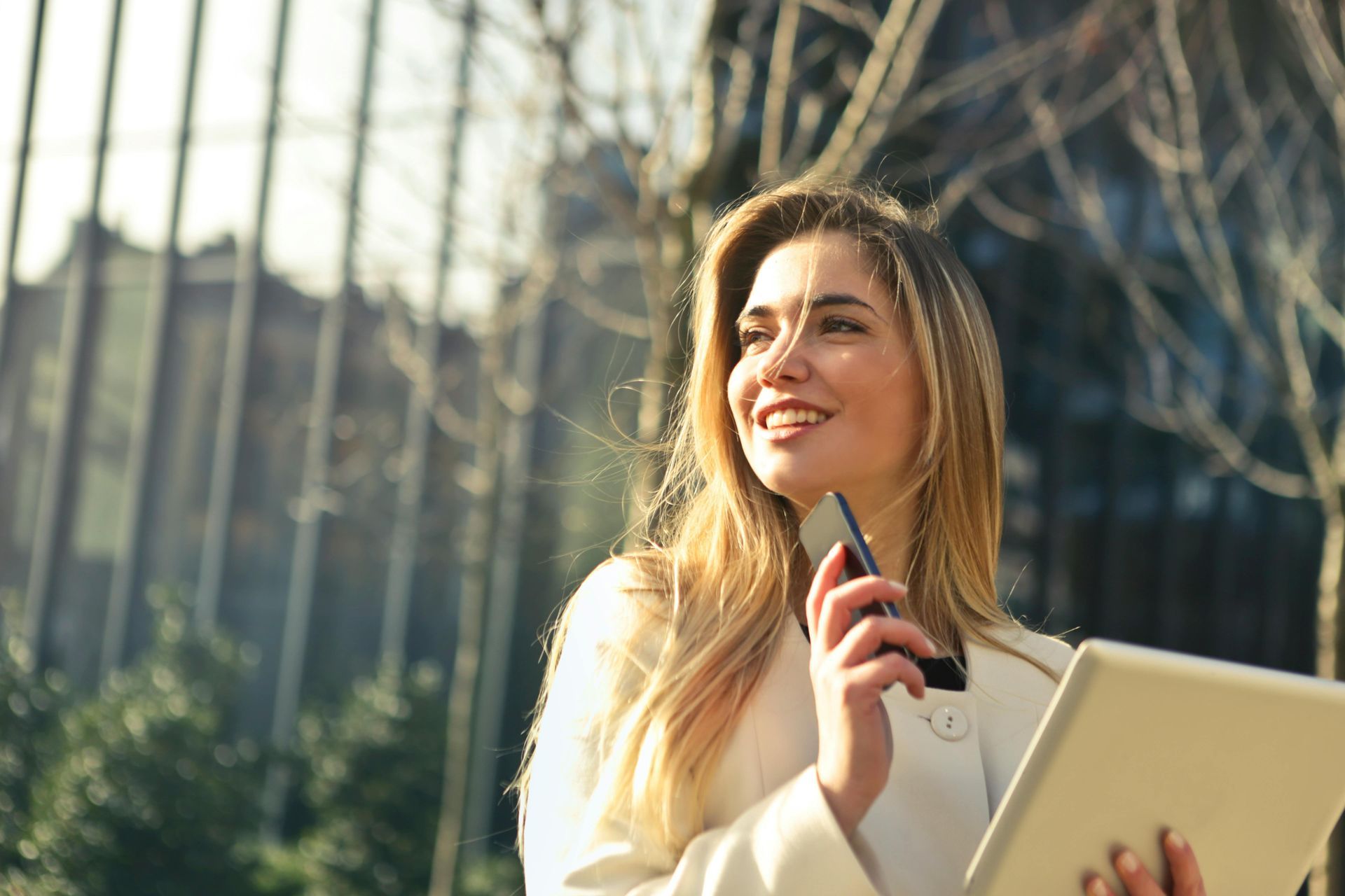 Woman smiling while holding a phone and laptop outdoors near glass buildings