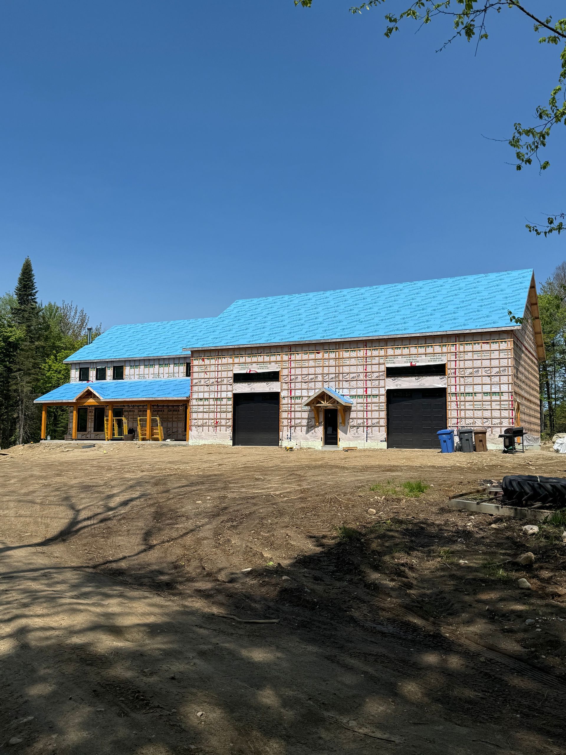 Une maison en construction, avec un bardage clair, une sous-toiture bleue et deux portes de garage noires, dans une zone boisée.
