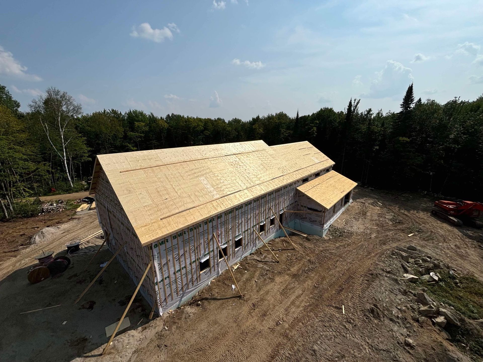 Vue aérienne d'une maison partiellement construite, avec une toiture en bois et des murs revêtus, sur un terrain en terre battue près d'une forêt.