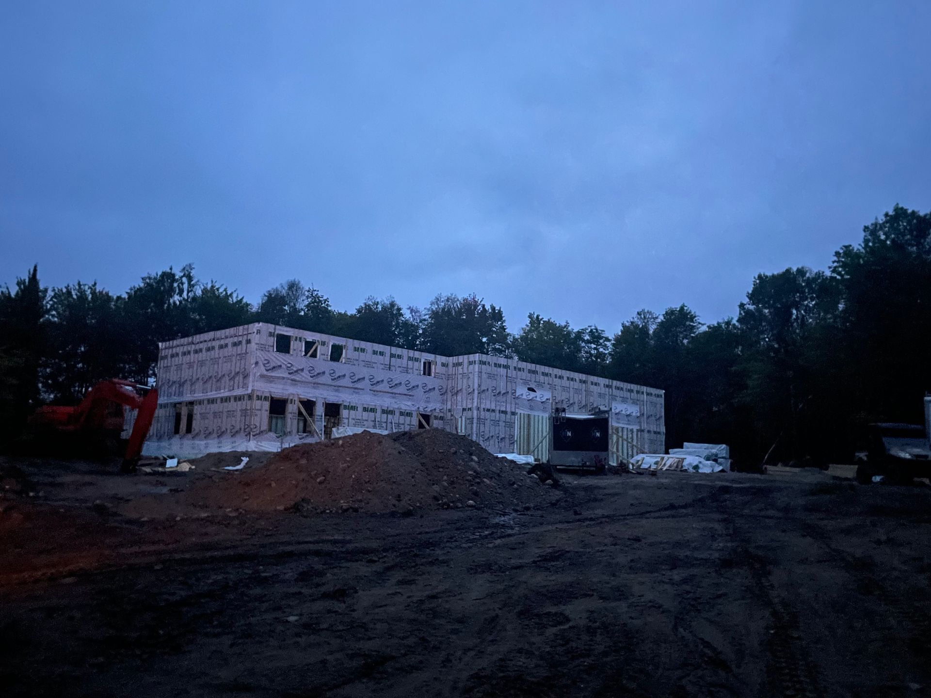 Un bâtiment en construction, au revêtement clair, se dresse sur un terrain vague sous un ciel sombre de crépuscule.