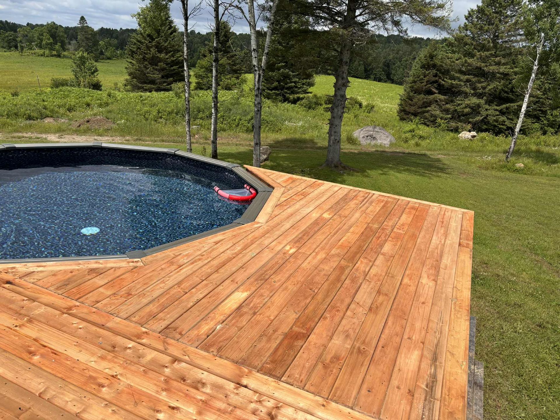 Une piscine ronde avec un revêtement bleu foncé, entourée d'une terrasse en bois fraîchement construite, dans un champ herbeux arboré.