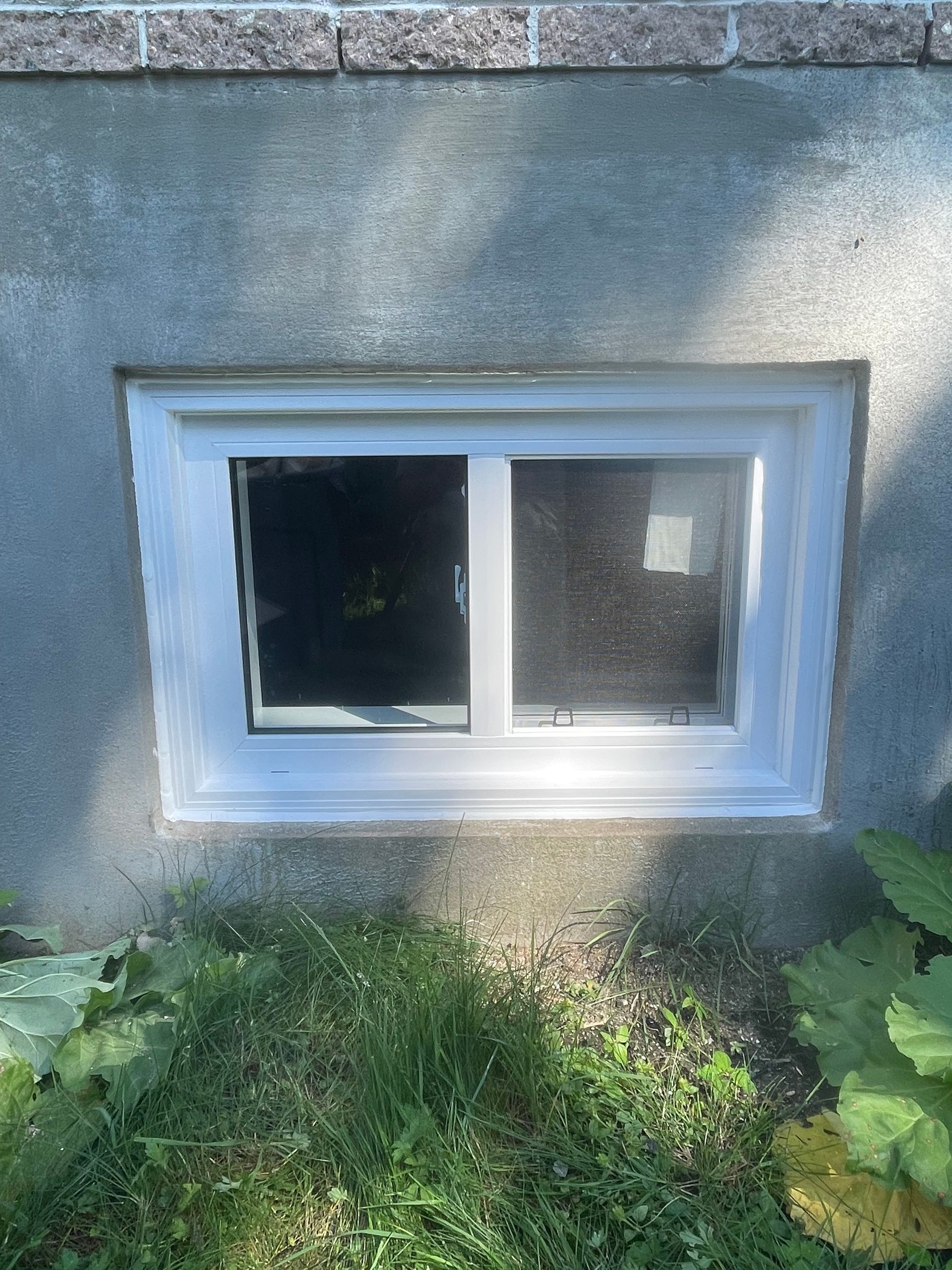 Une fenêtre coulissante à cadre blanc, installée dans un mur de fondation en béton gris, au-dessus d'un coin de terre et de verdure.