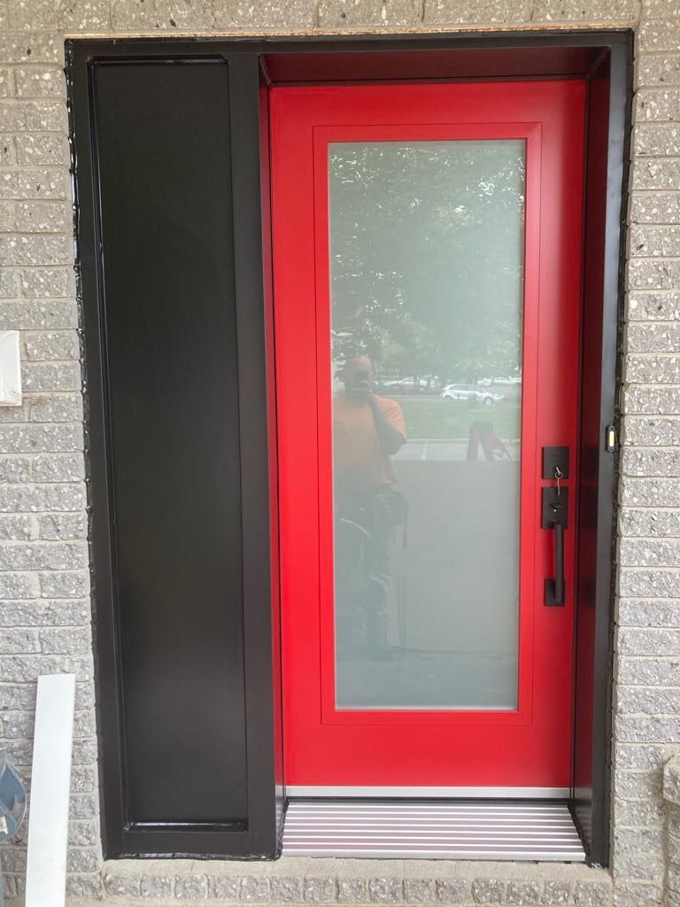 Une porte d'entrée moderne rouge vif avec un panneau vertical en verre dépoli, installée dans un cadre sombre contre un mur de briques claires.