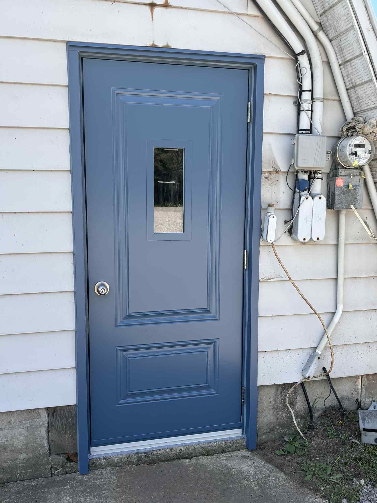 Une porte rectangulaire bleu foncé avec une petite fenêtre centrale, installée dans un bâtiment aux murs blancs, à côté d'équipements techniques.