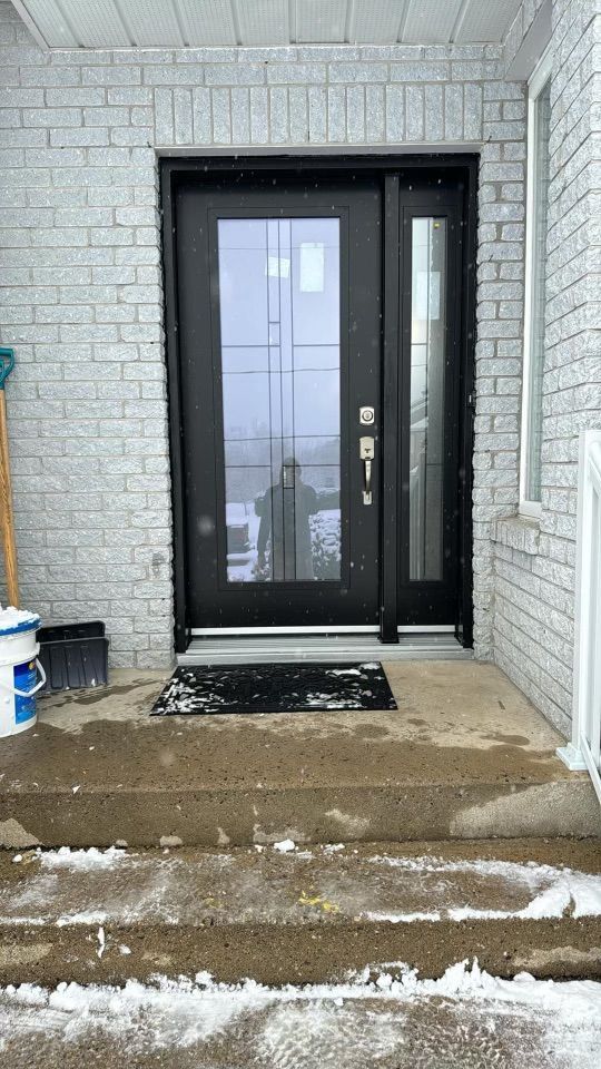 Une porte d'entrée moderne noire avec un panneau de verre et une imposte latérale, encastrée dans un mur de briques gris clair, avec de la neige sur les marches en béton.