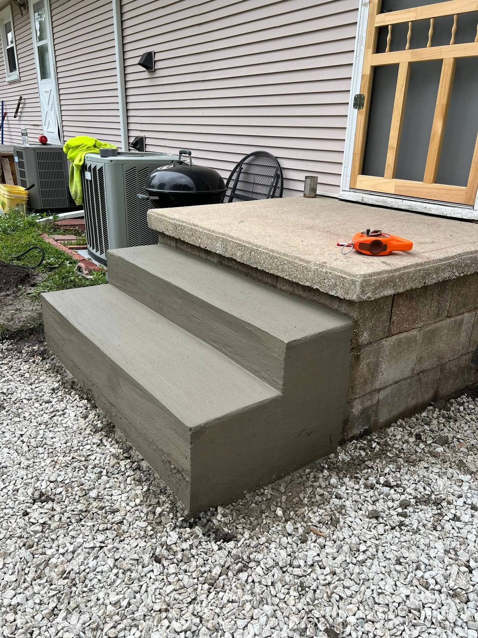 Concrete steps leading up to a raised patio with a grill. Exterior shot with gravel.