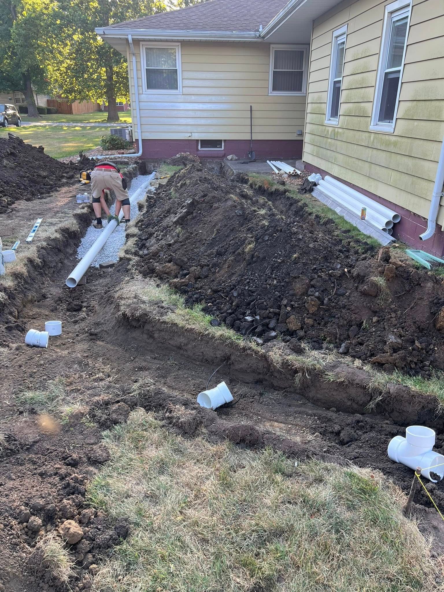 A person installing drainage pipes near a house. Trenches, dirt piles, and white PVC pipes are visible.