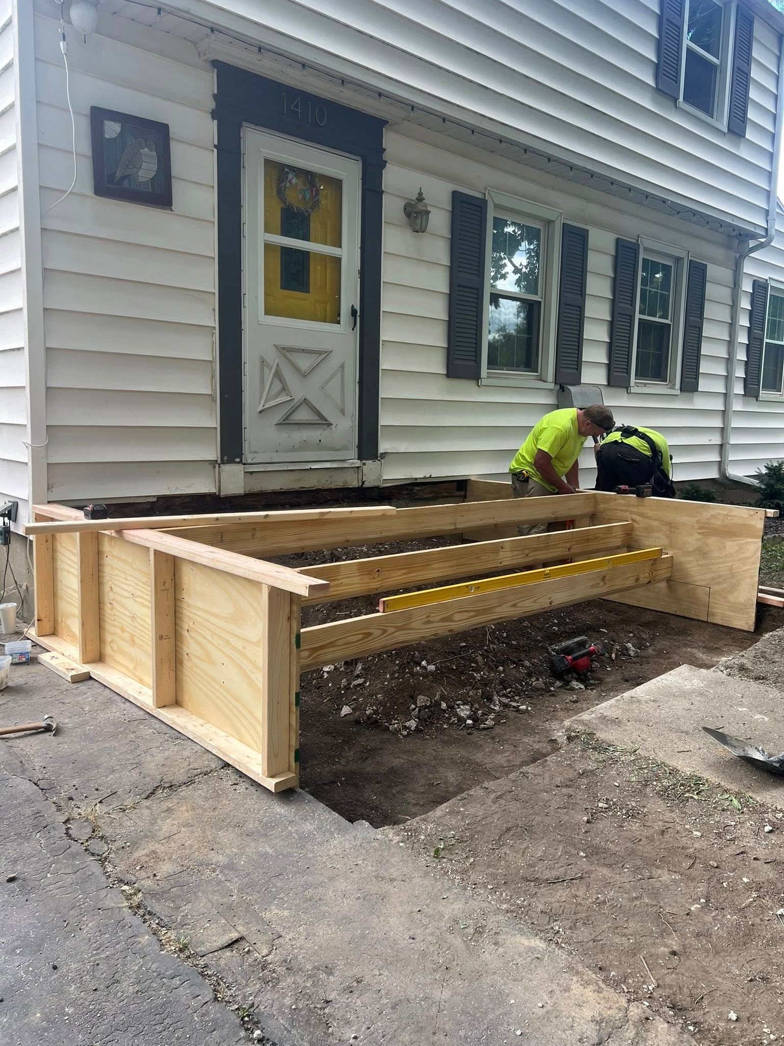 Construction of wooden steps in front of a white house. Two workers are present.