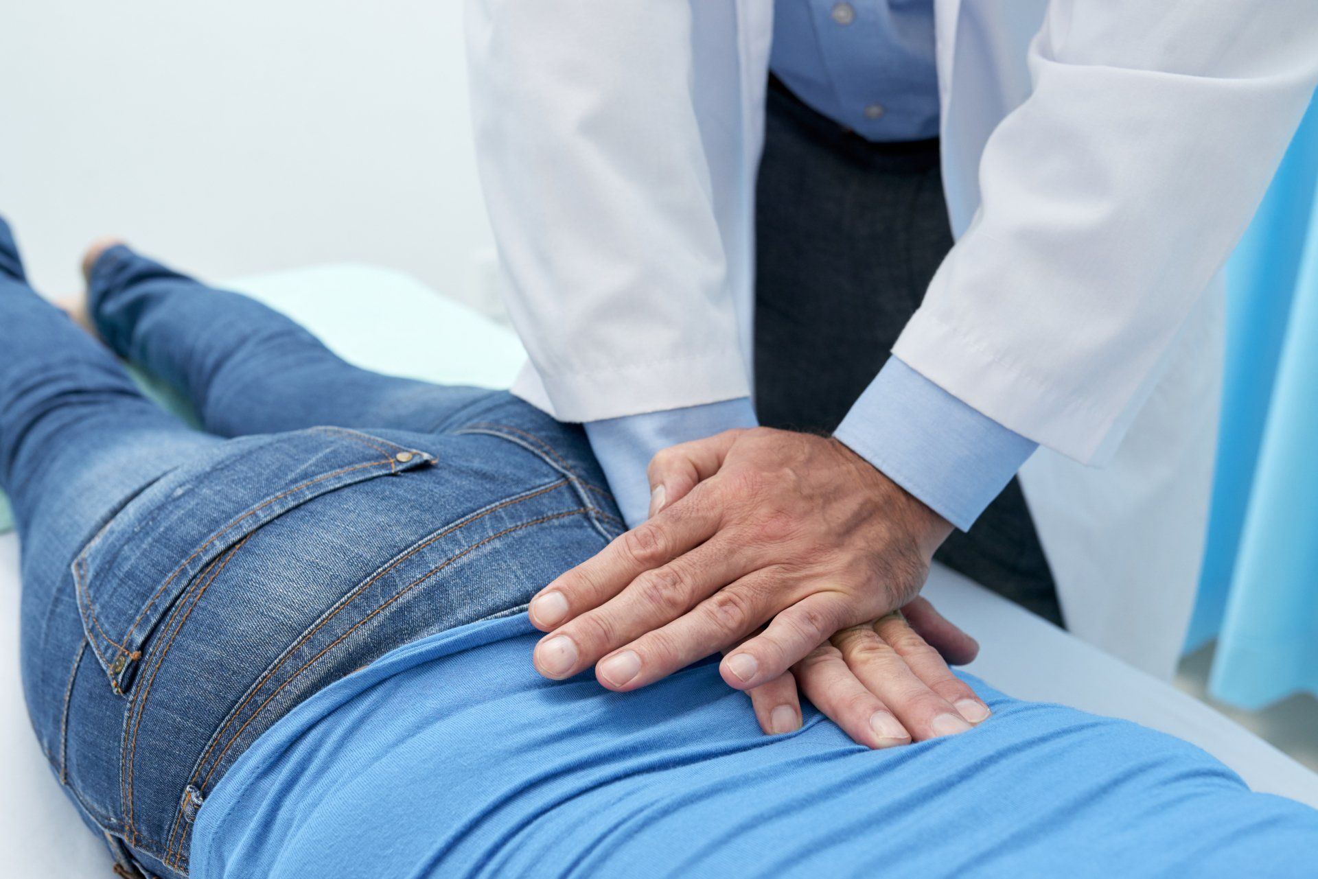 Doctor examining a patient's lower back. Blue shirt, jeans, and white coat in a medical setting.