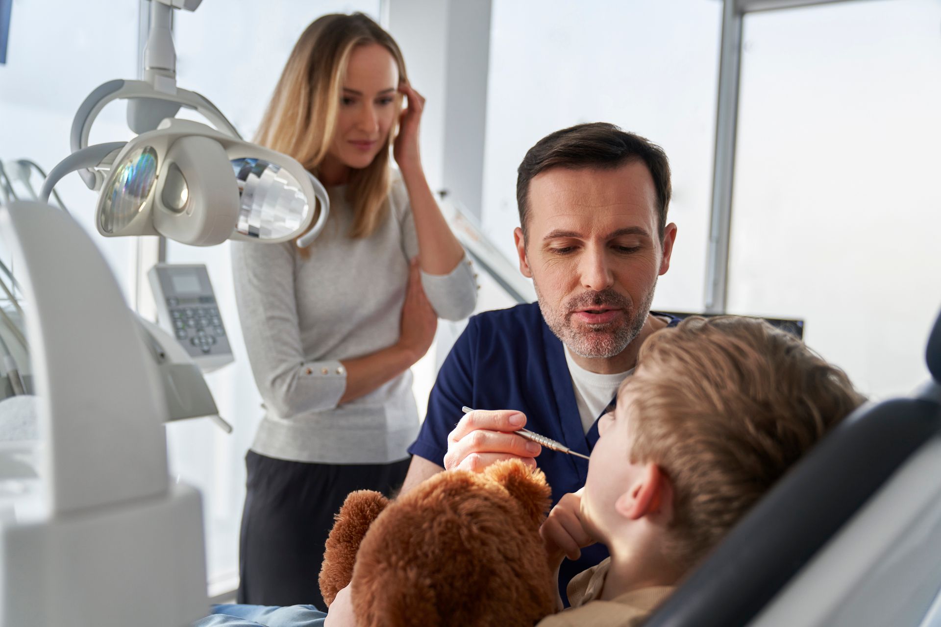 Little boy holding a teddy bear, while visiting a family dentist with his mother.