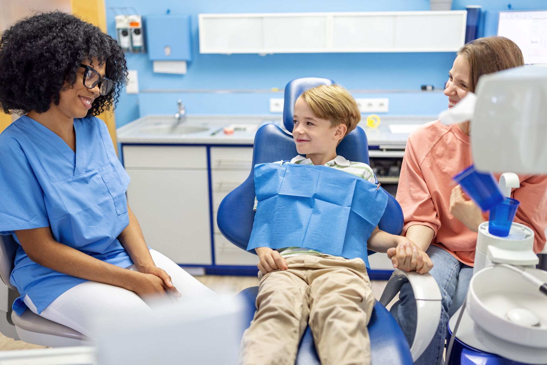 A boy is sitting in a dentist’s chair holding his mother's hand, smiling at the female dentist.