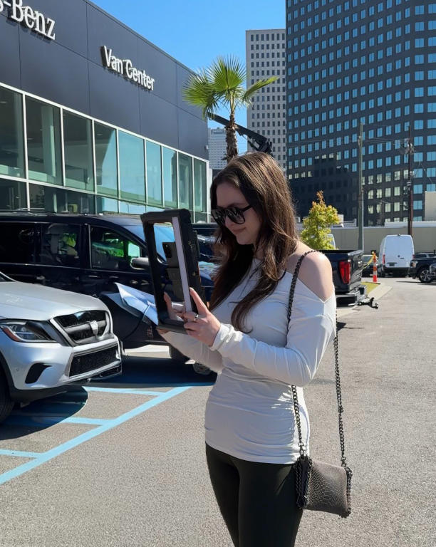 Woman in sunglasses looking at item in a frame in front of a Mercedes-Benz dealership.