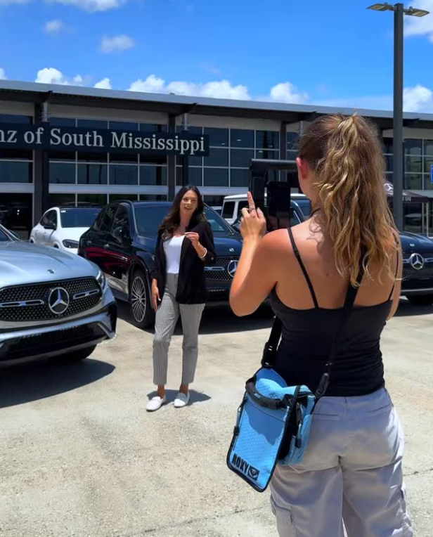 Woman being filmed in front of a Mercedes-Benz dealership. Cars, sunny outdoor setting.