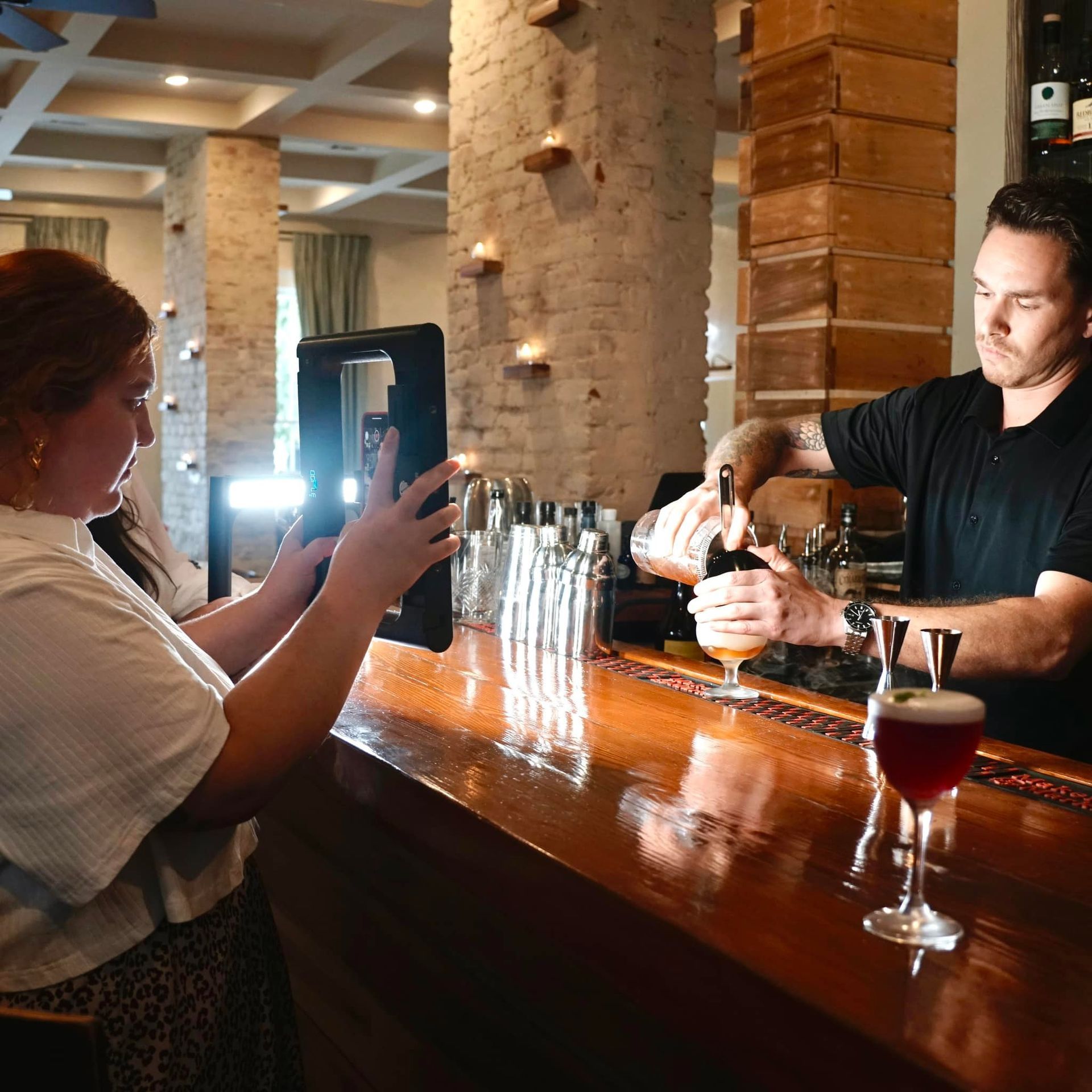 Woman filming bartender pouring drink at a bar.