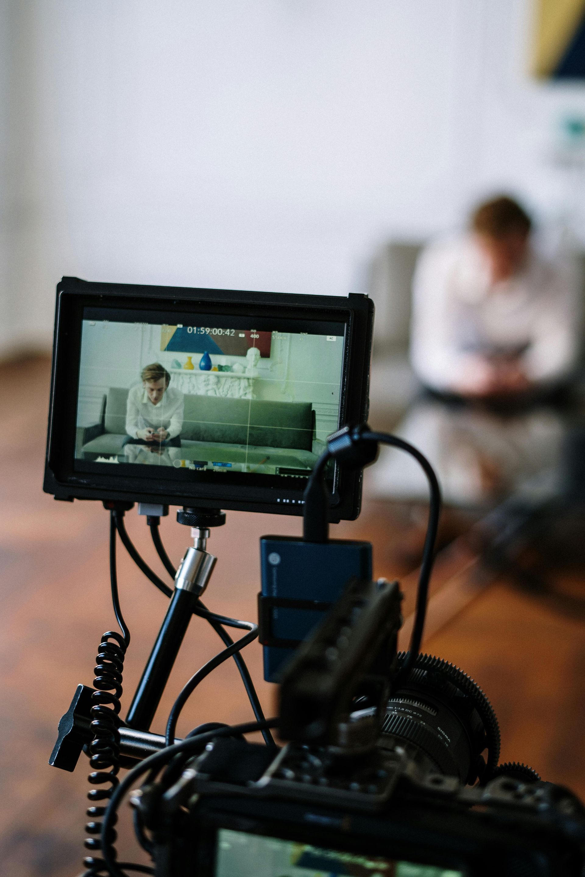 Camera setup filming a person indoors. Monitor shows person on sofa, camera and wires in foreground.