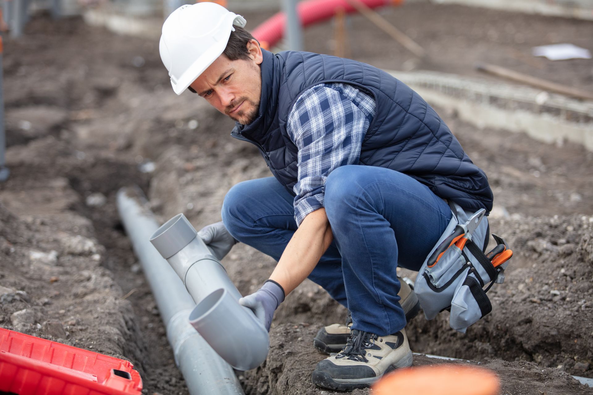 Construction worker in hard hat crouching beside gray pipes at a muddy roadside worksite.