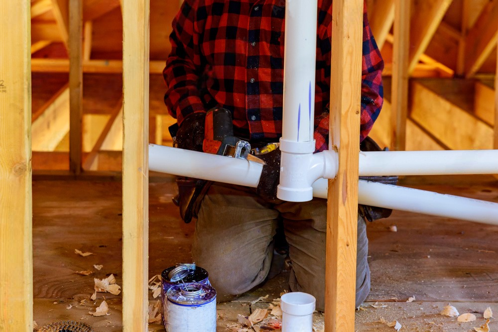 Plumber installing white PVC pipes under a wooden house floor, wearing a red plaid shirt and tan pants