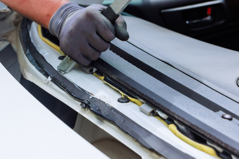 Gloved hand using a tool to repair the edge of a white car windshield