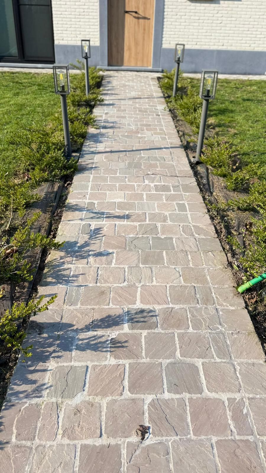 Een stenen pad leidt naar een houten voordeur, omlijst door groene struiken en op een zonnige dag verlicht door vier moderne padverlichting.