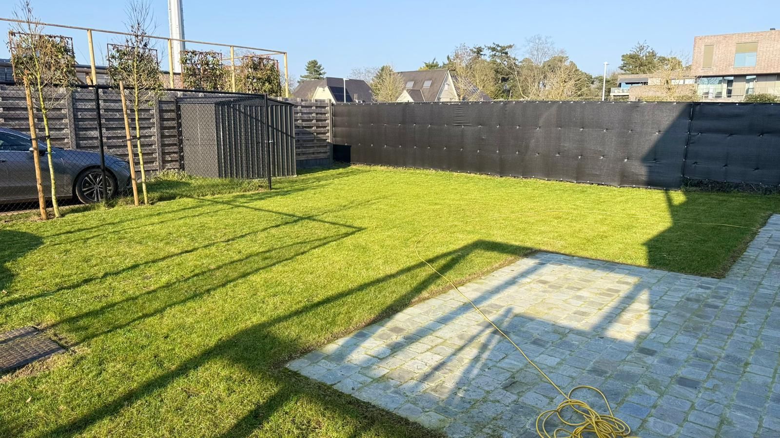 Een zonnige achtertuin met een betegeld terras, een gazon, een auto geparkeerd bij een hek en een zwart privacyscherm.