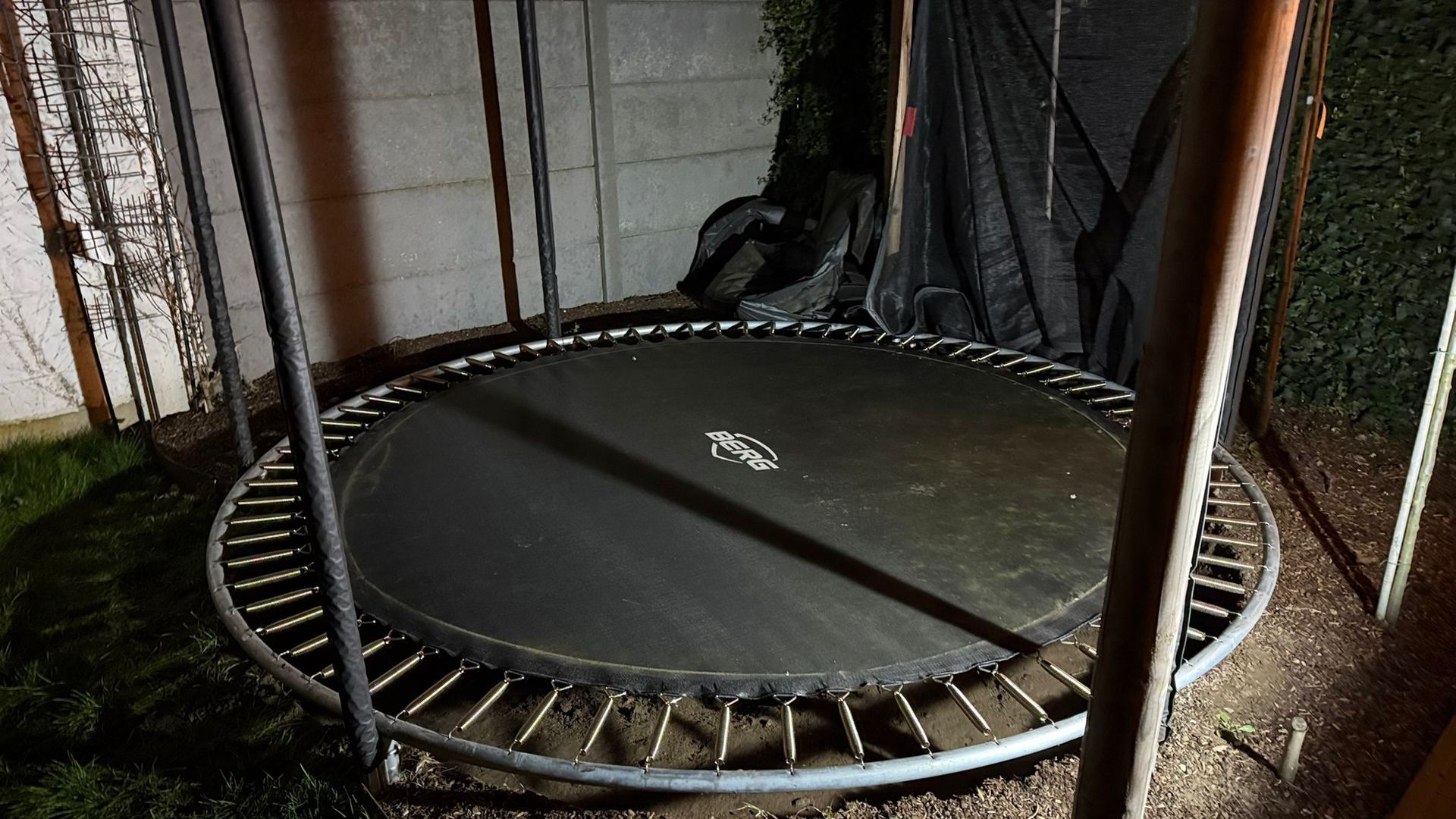 Een zwarte, ronde trampoline met een zichtbaar metalen frame, buiten geplaatst in een schemerig verlichte ruimte vlakbij een muur.