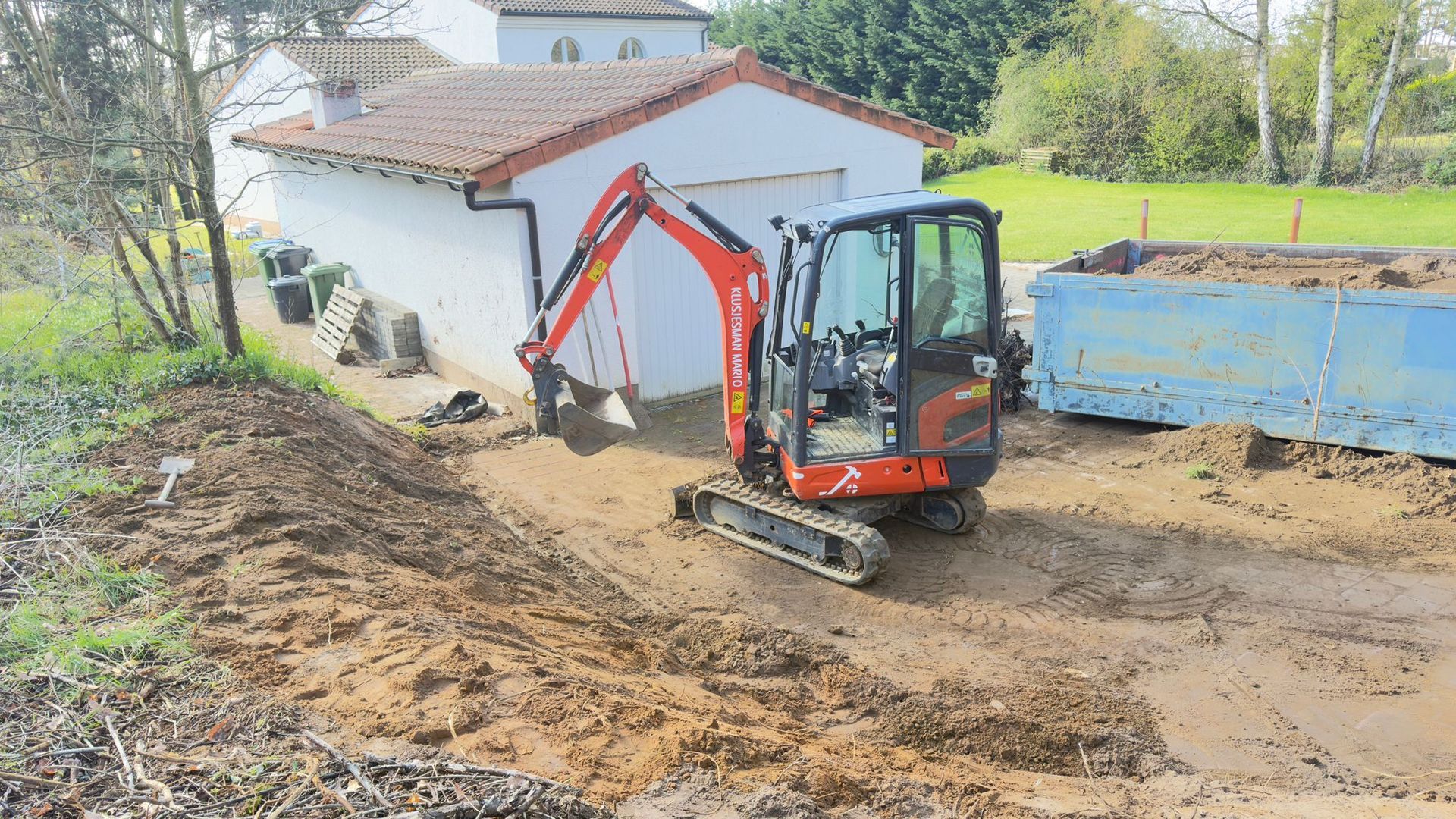 Een oranje compacte graafmachine staat op een modderig werkterrein naast een wit gebouw en een grote blauwe afvalcontainer.
