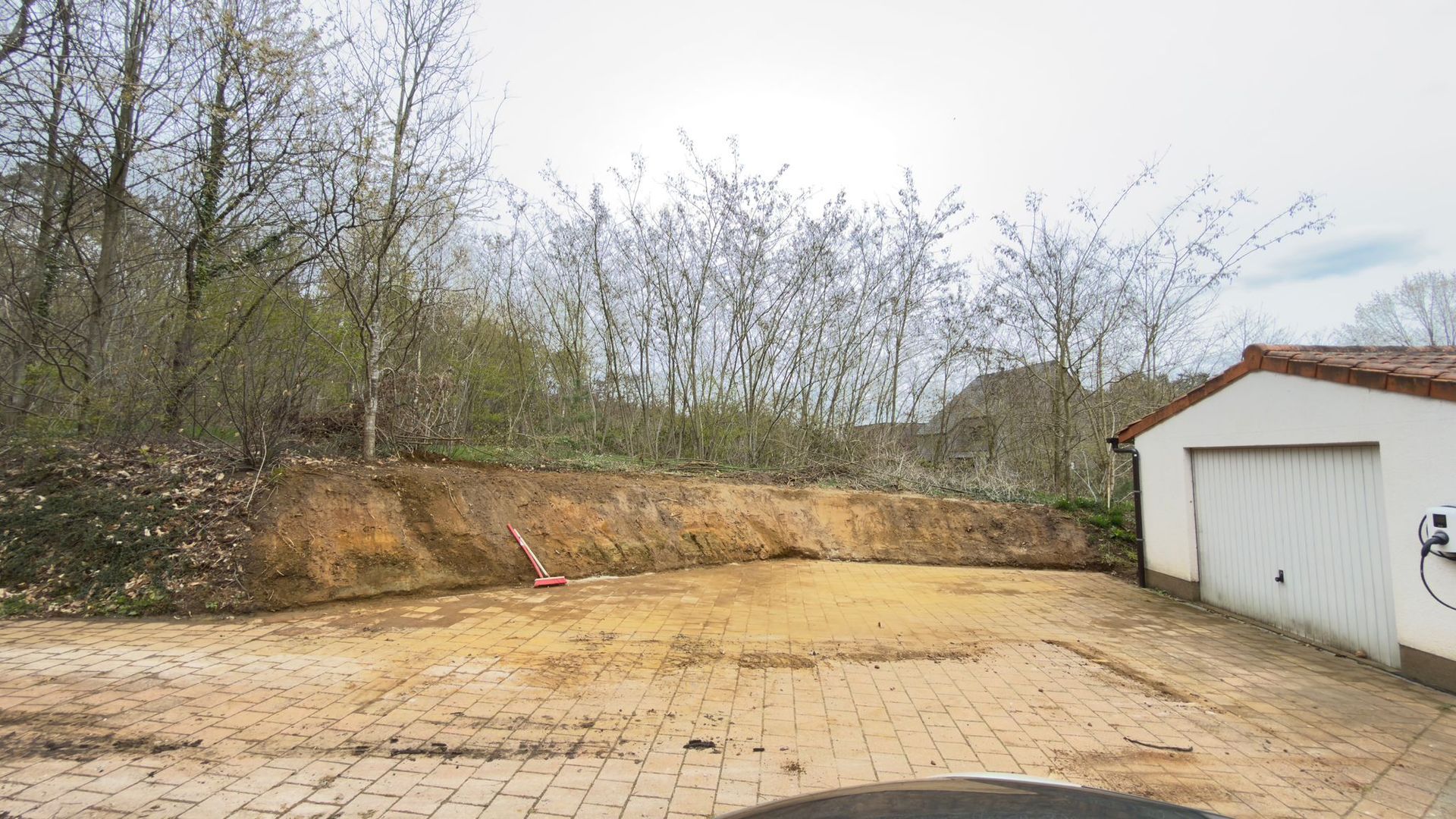 Een grindterrein naast een garage met een lage aarden wal, begrensd door kale bomen tegen een bewolkte hemel.